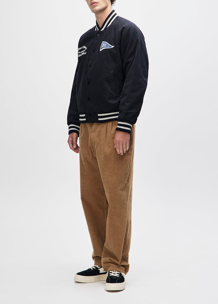 Person wearing a black varsity jacket with white stripes, brown pants, and black sneakers on a white background
