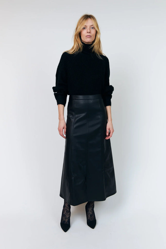 A woman stands against a plain light gray studio background wearing a black ribbed turtleneck sweater, a black leather-look midi skirt, black lace tights, and black pointed heels.