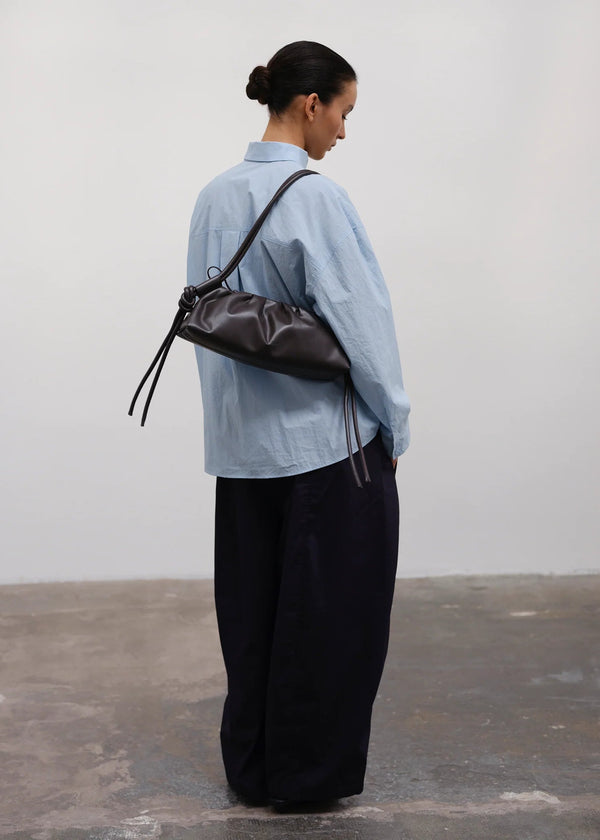 Person wearing a light blue shirt and black pants, carrying a brown leather bag against a plain background.
