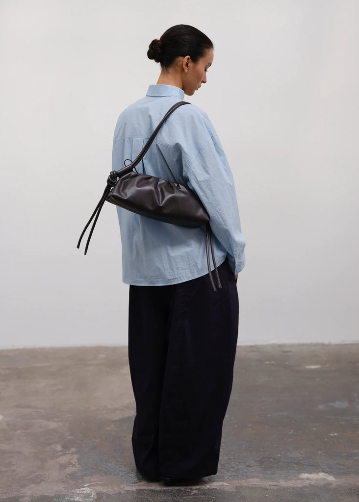 Person wearing a light blue shirt and black pants, carrying a brown leather bag against a plain background.