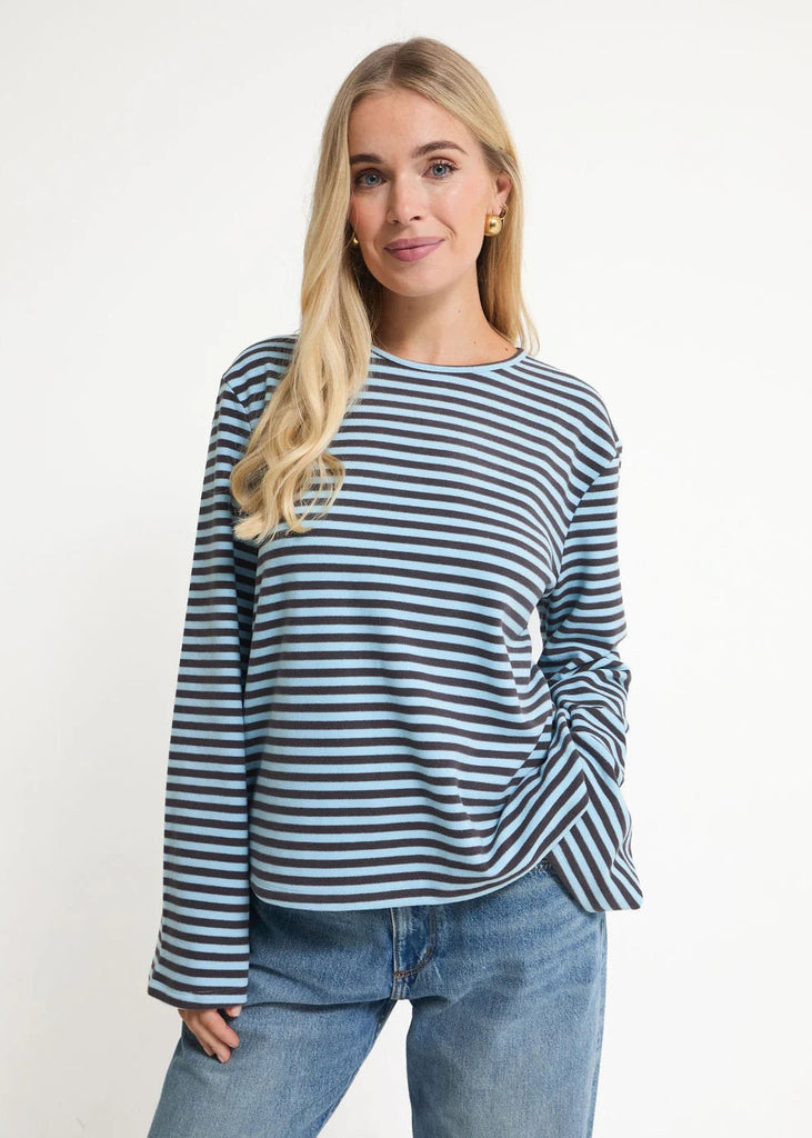 A woman modeling a long-sleeve crewneck top with navy and light blue horizontal stripes, paired with light-wash blue jeans, against a plain white studio background.