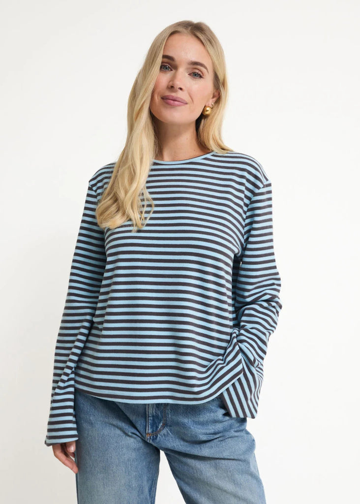 A woman modeling a long-sleeve crewneck top with horizontal navy and light blue stripes, paired with blue jeans, against a plain white studio background.