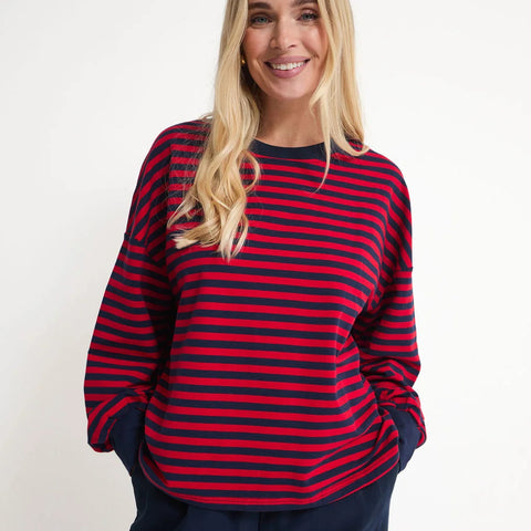 A smiling blonde woman wearing a red and navy blue horizontally striped long-sleeve top with navy pants, standing against a plain light gray studio background.