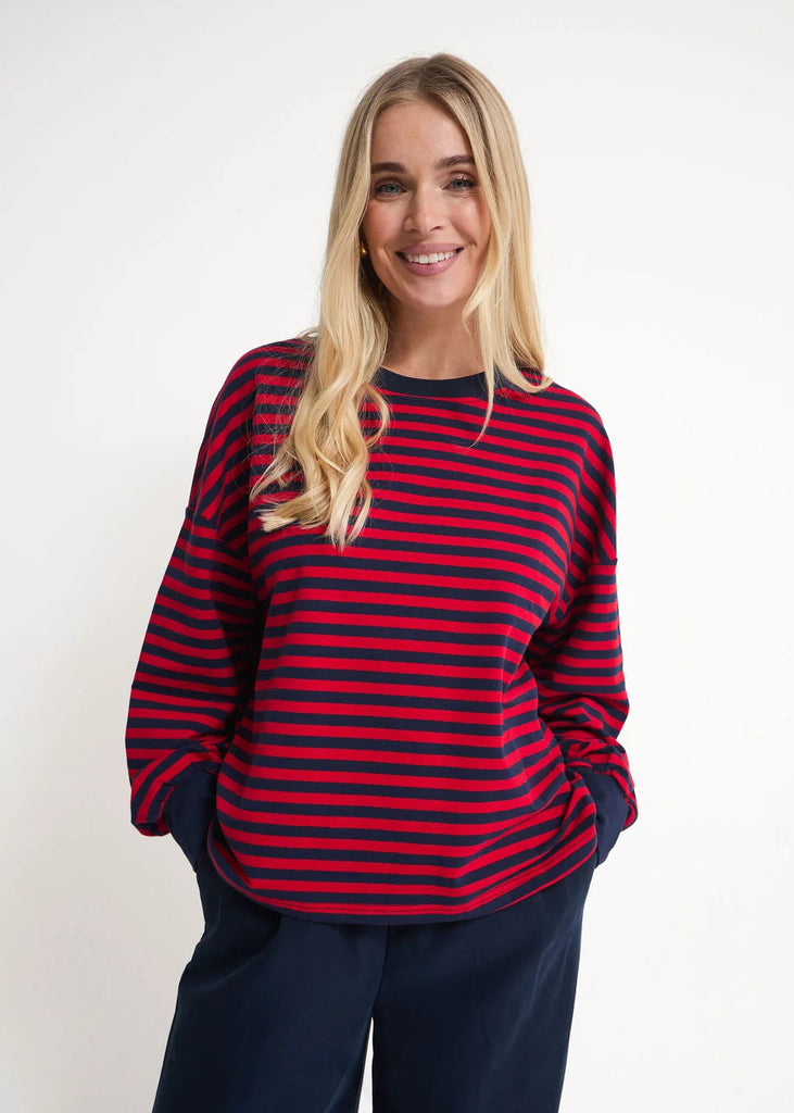 A smiling blonde woman wearing a red and navy blue horizontally striped long-sleeve top with navy pants, standing against a plain light gray studio background.