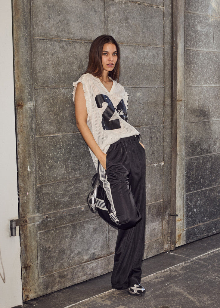 Woman wearing a white sleeveless top with the number 24 and black pants, leaning against a textured wall.