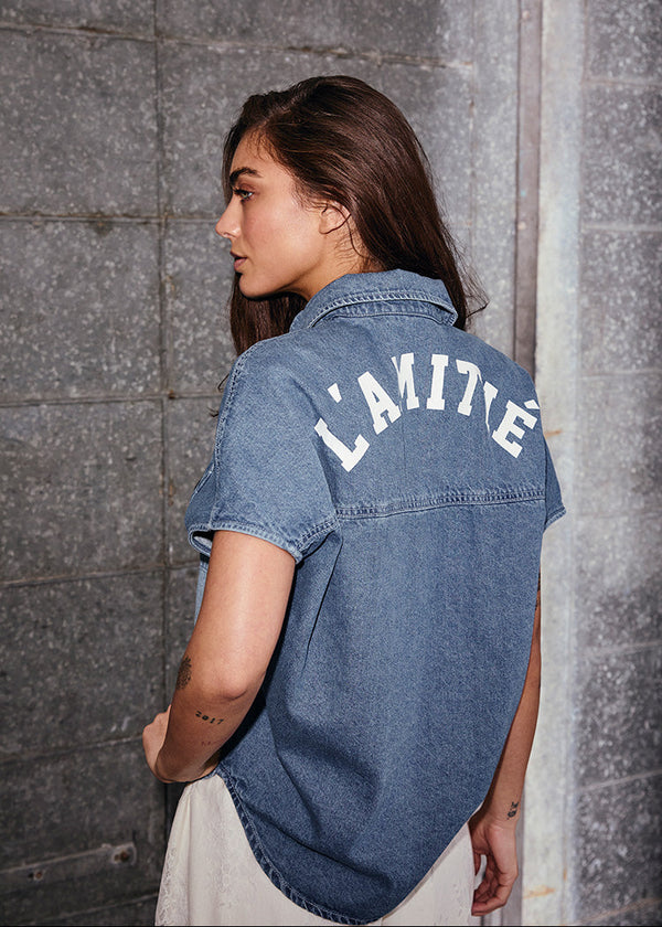 Woman wearing a blue denim shirt with 'L'AMOUR' text against a gray wall.