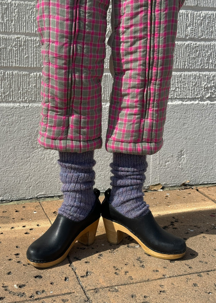 Person wearing pink plaid pants, black clogs, and purple socks standing on a sidewalk.