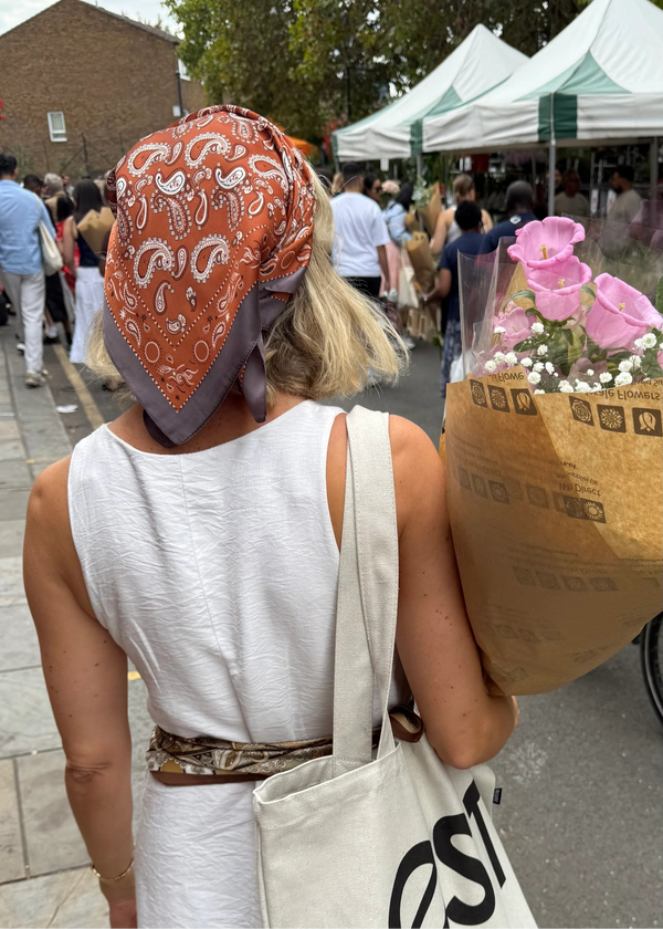 Woman with orange headscarf and white tote bag walking outdoors with a crowd and tents in the background