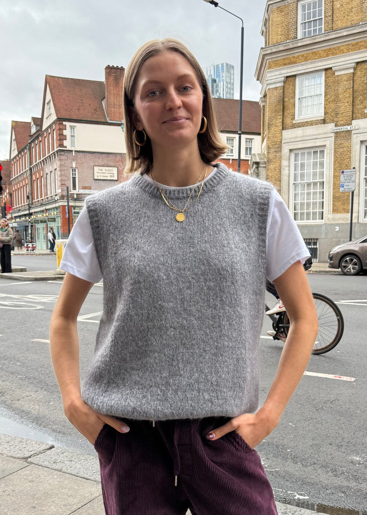 Person wearing a gray sleeveless sweater and purple pants standing on a city street.