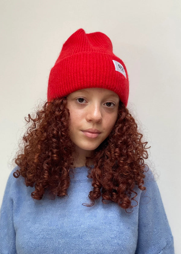 A person with curly red hair wearing a bright red knit beanie and a light blue sweater, photographed against a plain light background.