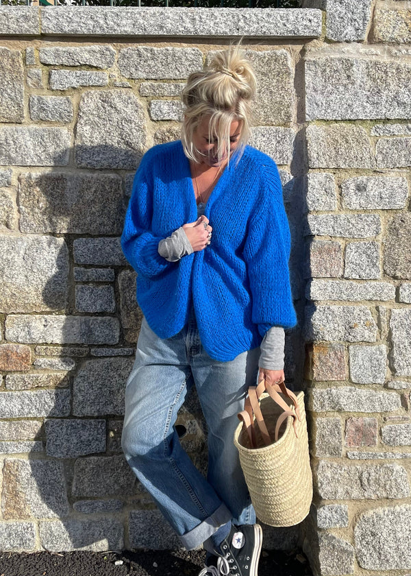 A person wearing a cobalt blue cardigan with a relaxed silhouette, paired with jeans, standing with one foot on a stone surface, holding a wicker tote bag.