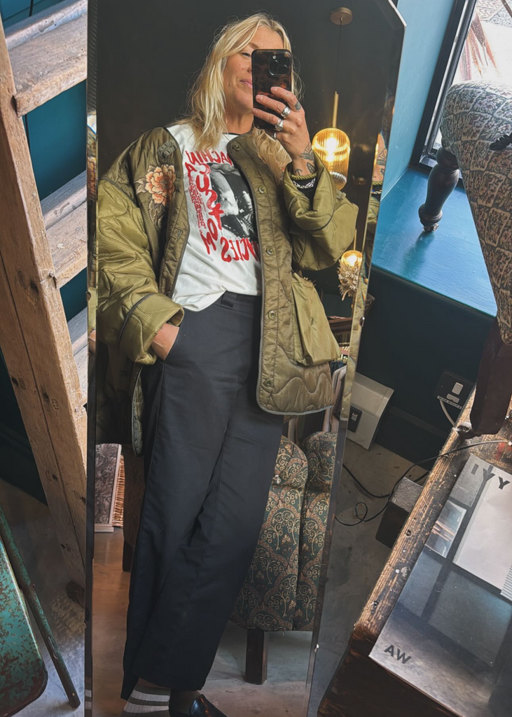 Person taking a mirror selfie wearing a green jacket and dark pants in an indoor setting.