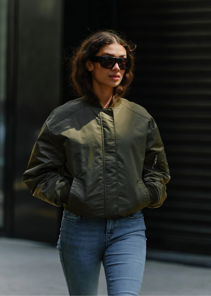 Woman wearing a green bomber jacket and blue jeans against a dark background