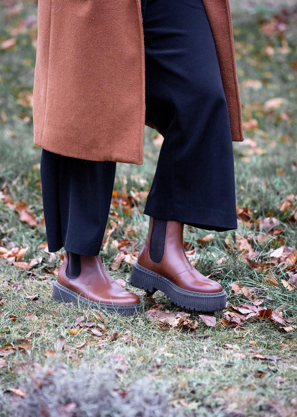 Person wearing brown leather boots and a long coat standing on grass with fallen leaves.