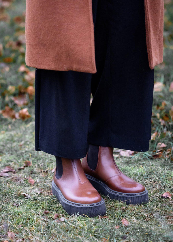 Brown leather boots worn with black pants and a brown coat on grass.