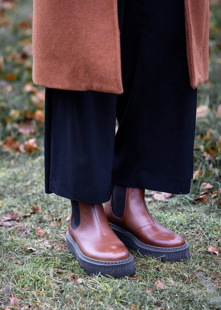 Brown leather boots worn with black pants and a brown coat on grass.