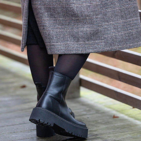 Person wearing black boots and a gray coat on a wooden floor.