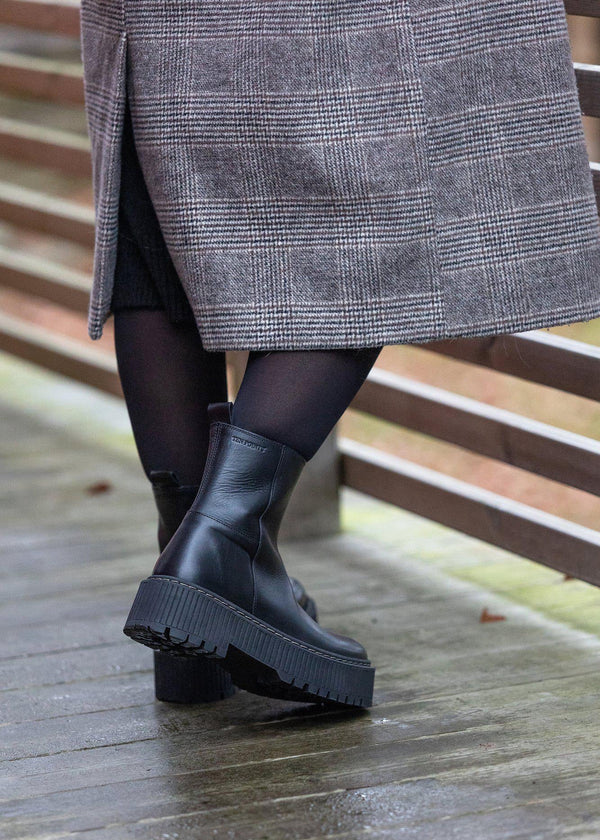 Person wearing black boots and a gray coat on a wooden floor.