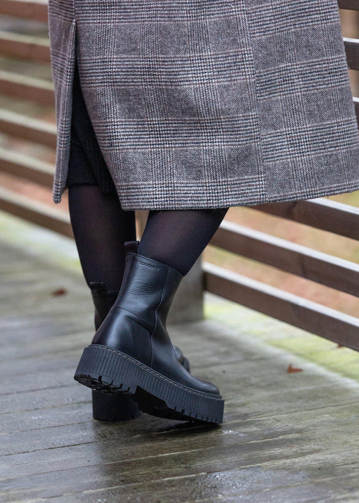Person wearing black boots and a gray coat on a wooden floor.