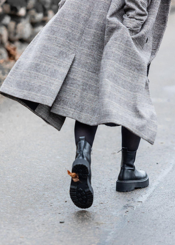Person wearing a long gray coat and black boots on a sidewalk.