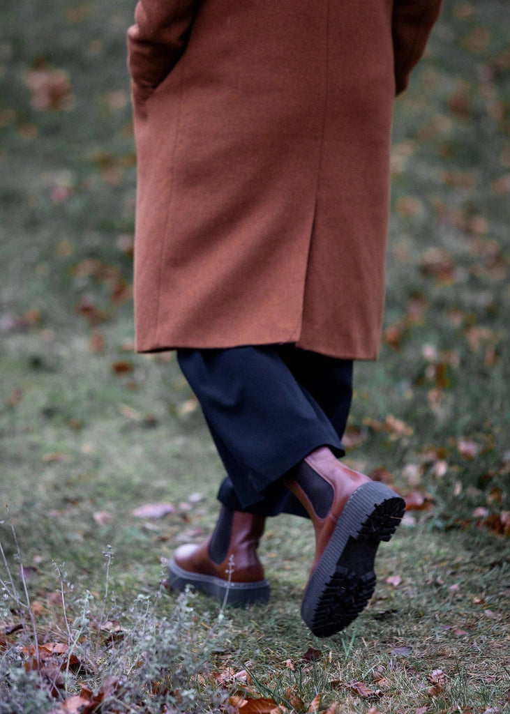 Person wearing a brown coat and dark boots walking on grass with fallen leaves.