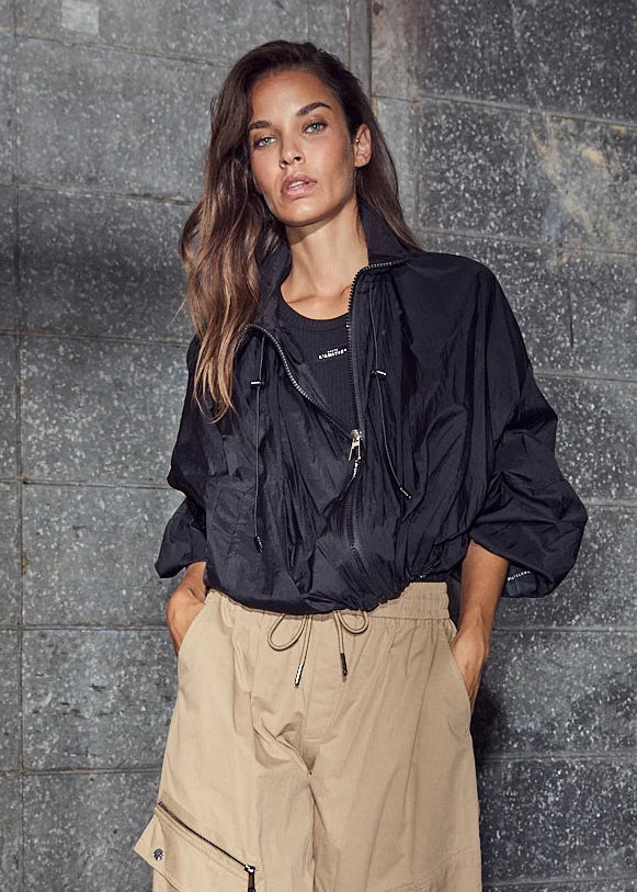 A woman stands against a dark gray concrete wall wearing baggy beige cargo pants, a black jacket, and white heeled sandals.