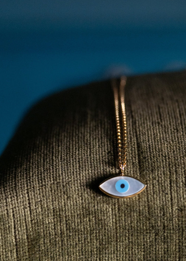 A gold necklace with a blue and white evil eye pendant resting on an antique gold wooden bench with a dark green cushion, set against a dark blue background.