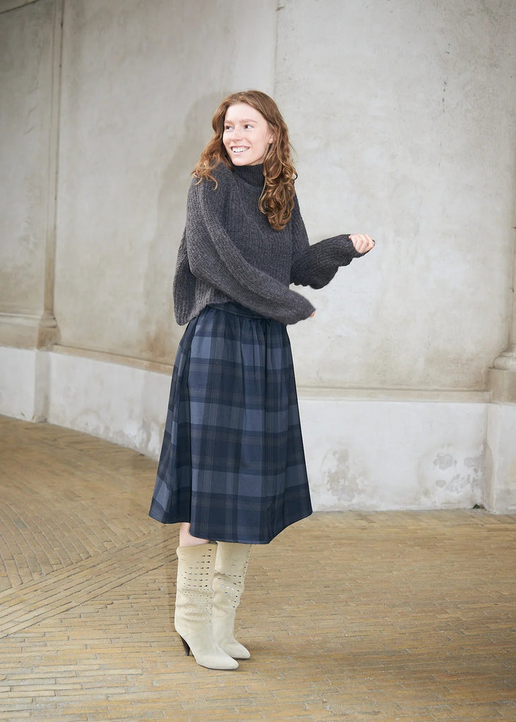 A woman with long wavy light brown hair stands in a real architectural setting with a textured plaster wall and tiled floor, wearing a dark gray fuzzy turtleneck sweater, a navy and gray plaid midi skirt, and off-white knee-high boots.