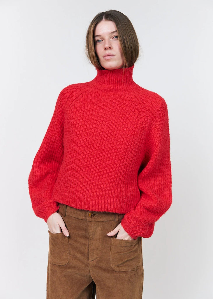 A woman wearing a bright red ribbed knit turtleneck sweater and brown corduroy pants, standing against a light gray studio background.