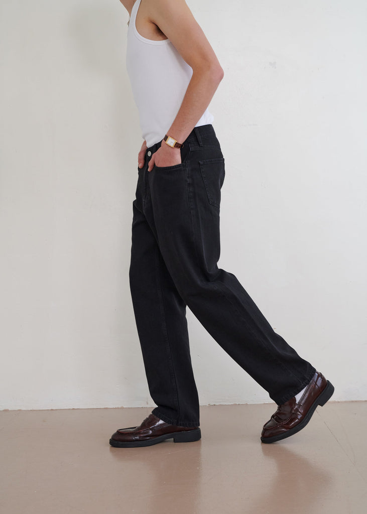 Person wearing a white tank top, black pants, and brown shoes against a plain background
