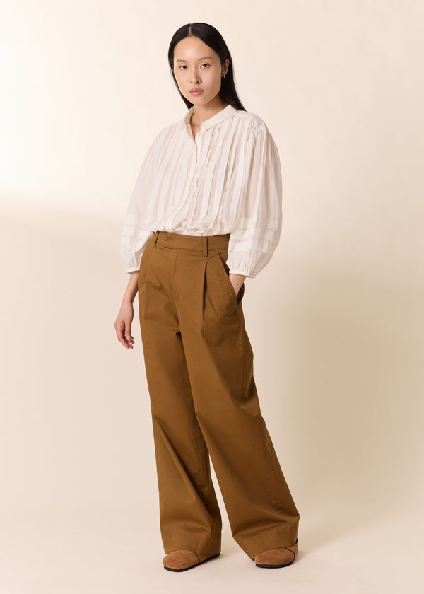 Woman wearing a white blouse and brown pants against a beige background