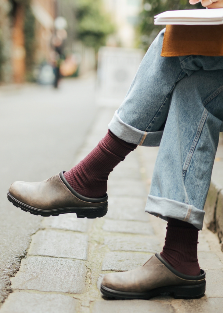Person sitting on a street curb wearing brown boots, rolled-up jeans, and maroon socks.