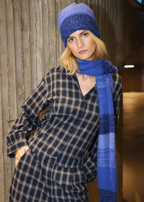 Woman wearing a blue knit hat and scarf with a plaid coat against a wooden wall.