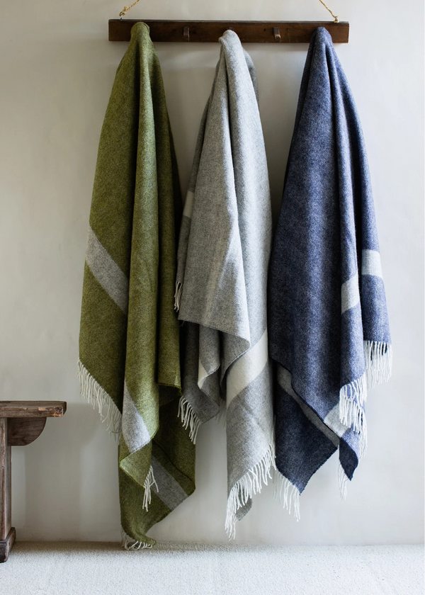 Three woven cloths in green, gray, and blue hanging on a wooden rod against a white wall.
