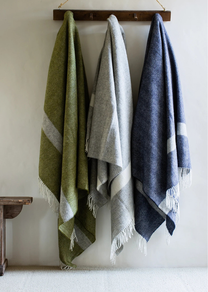 Three woven cloths in green, gray, and blue hanging on a wooden rod against a white wall.