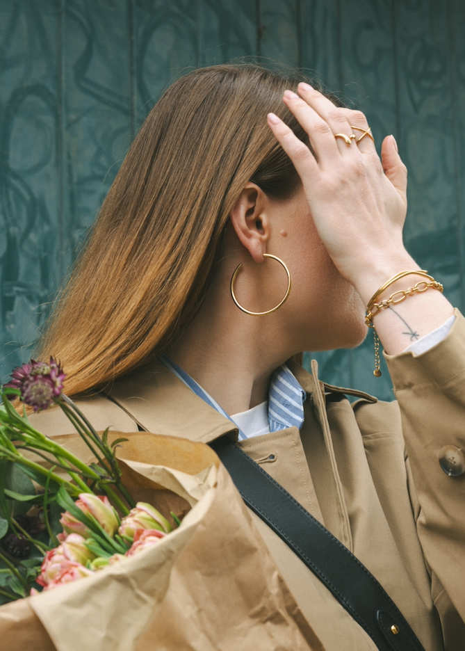 Woman in a beige coat holding flowers, wearing gold hoop earrings and bracelets, against a textured green wall.