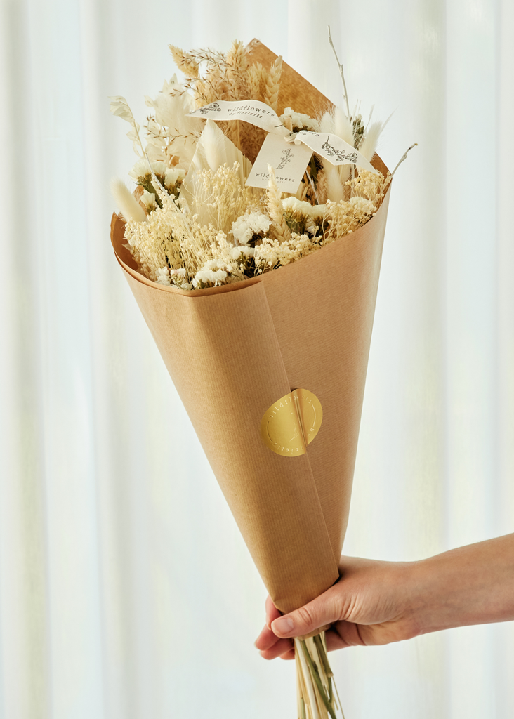 Bouquet of dried flowers wrapped in brown paper with a gold circle, held against a white curtain background.
