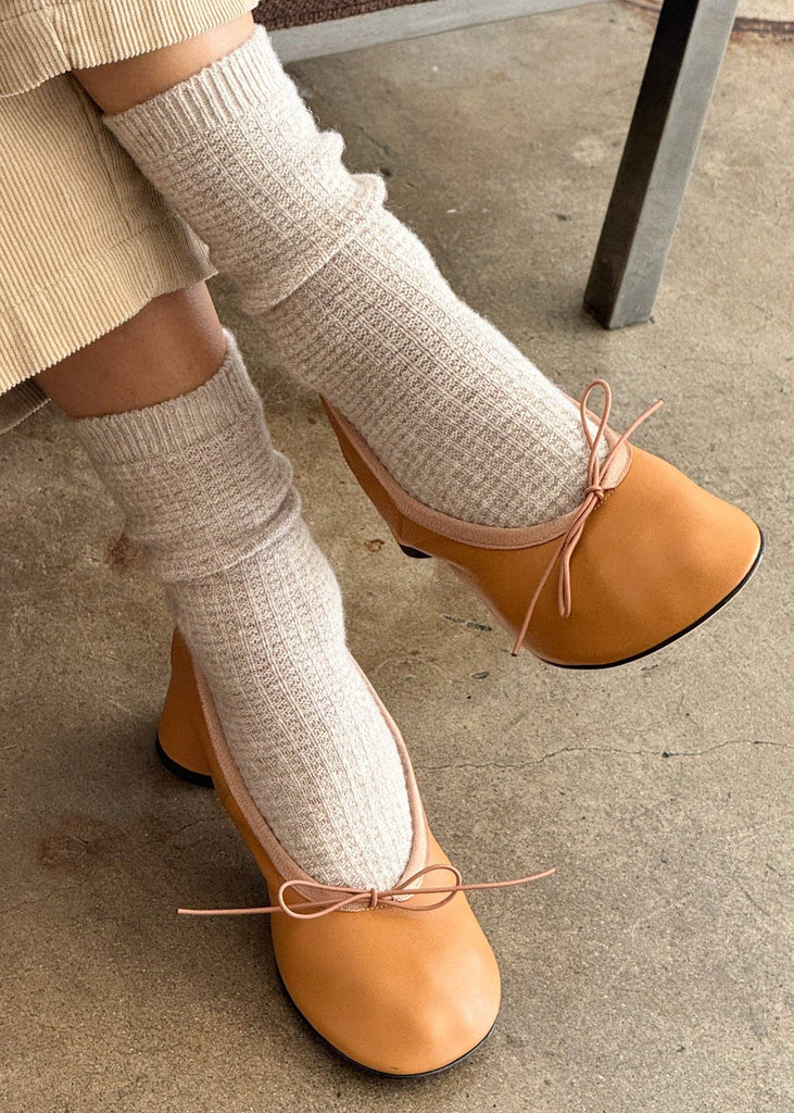 Person wearing beige shoes with bow details and white leg warmers on a concrete floor.