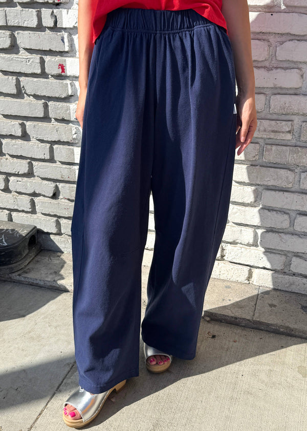 A person wearing loose, wide-leg navy pants with an elastic waistband, paired with a red top and metallic silver sandals, standing outdoors against a white brick wall.