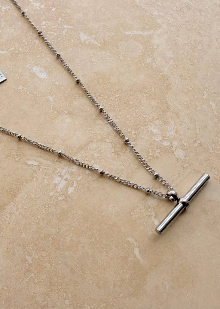 A silver-toned layered necklace with a beaded chain, a small engraved tag, and a polished T-bar clasp, displayed on a beige stone surface.