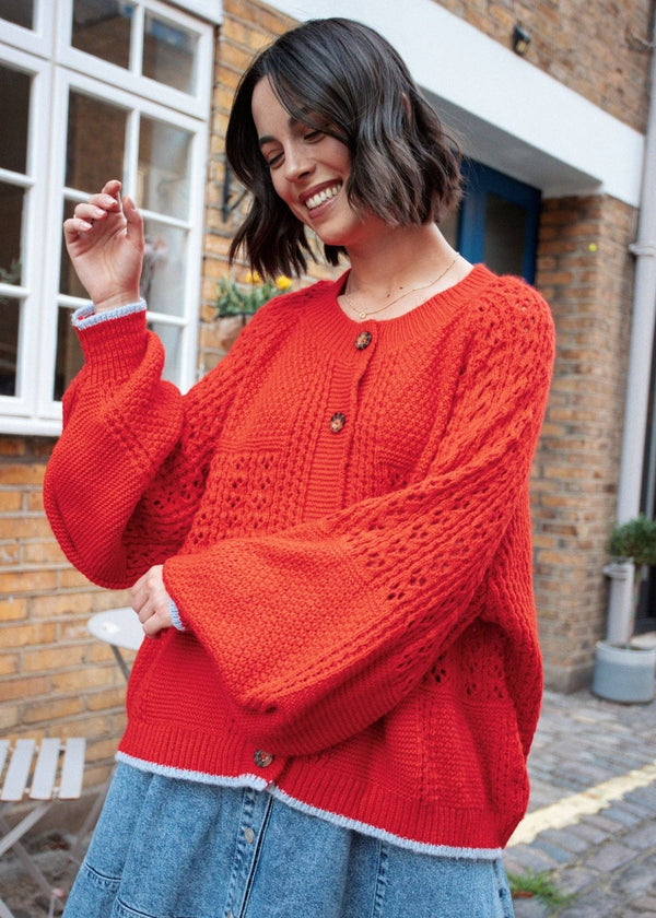 Woman wearing a red knitted sweater and blue jeans standing in an urban setting.