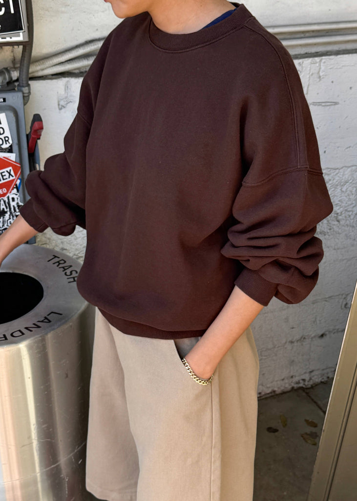 Person wearing a brown sweatshirt and beige pants standing near a trash can.