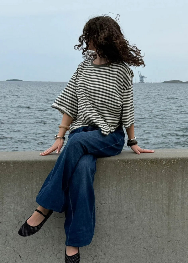 Person sitting on a concrete ledge by the water wearing a striped shirt and blue pants.