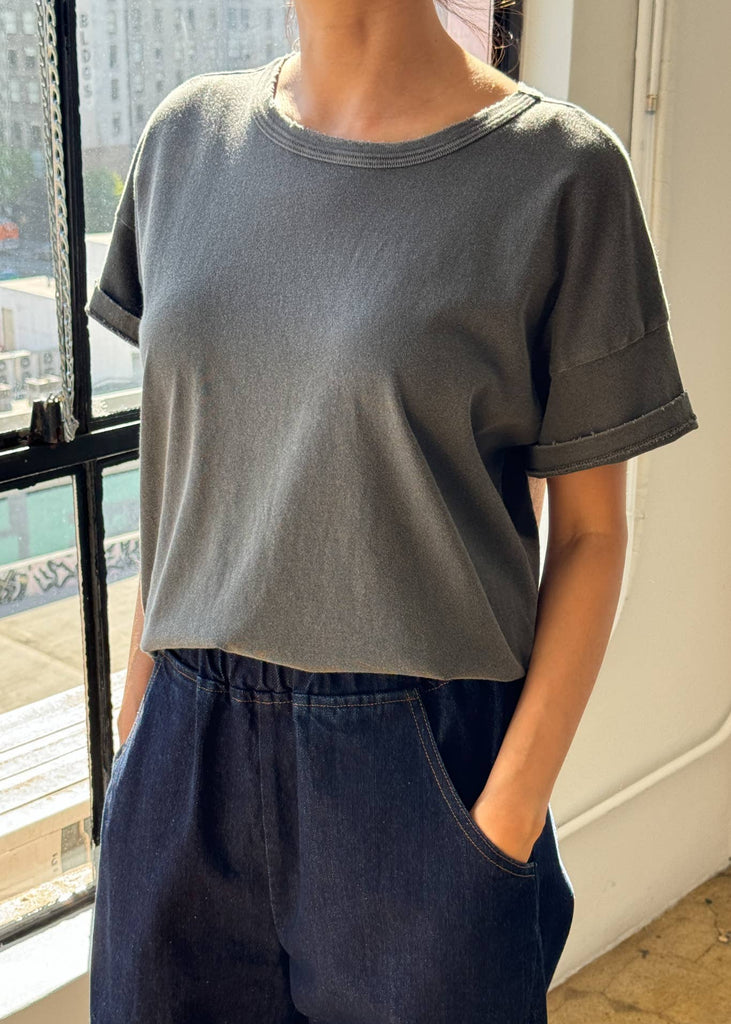 Person wearing a gray t-shirt and dark pants standing indoors with a window view.
