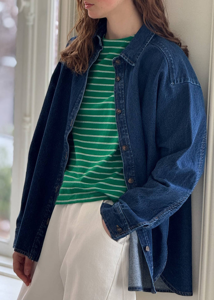Person wearing a green striped shirt, blue denim jacket, and white pants indoors.