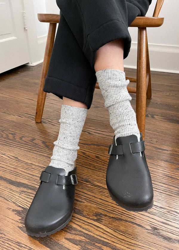 Person wearing black clogs and gray socks on a wooden floor.