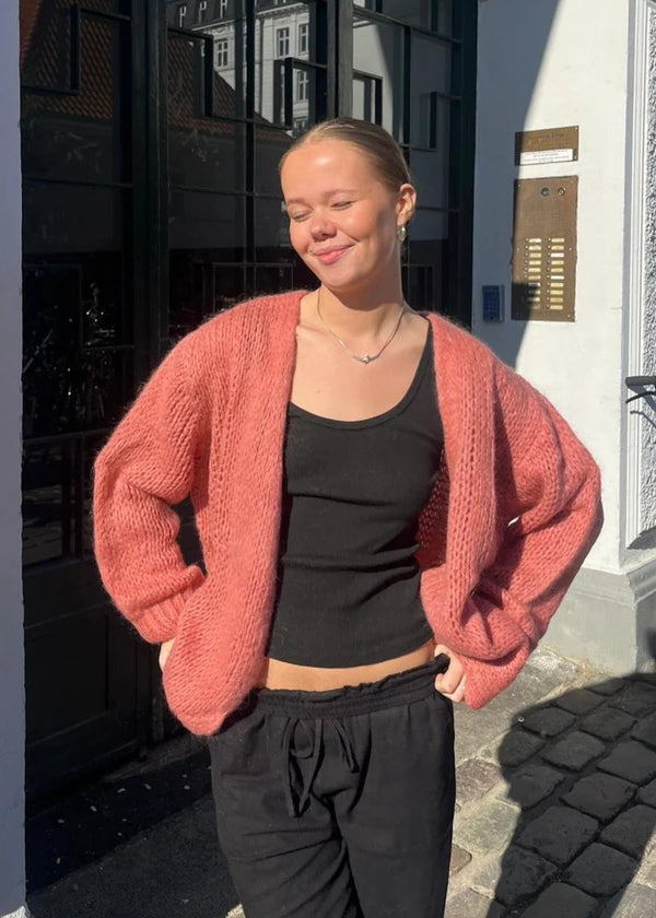 Person wearing a pink cardigan over a black top and pants, standing on a street.