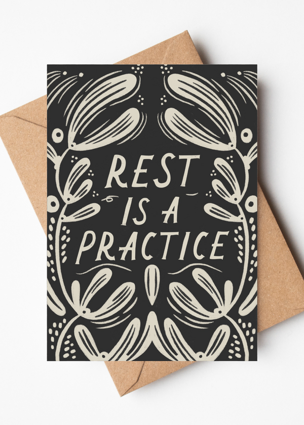 A rectangular greeting card with a black background and cream botanical illustration and the words “REST IS A PRACTICE,” shown with a kraft paper envelope on a light gray background.