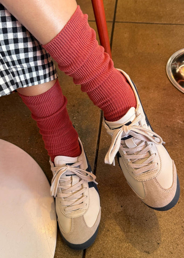 Person wearing red leg warmers and beige sneakers on a brown floor.