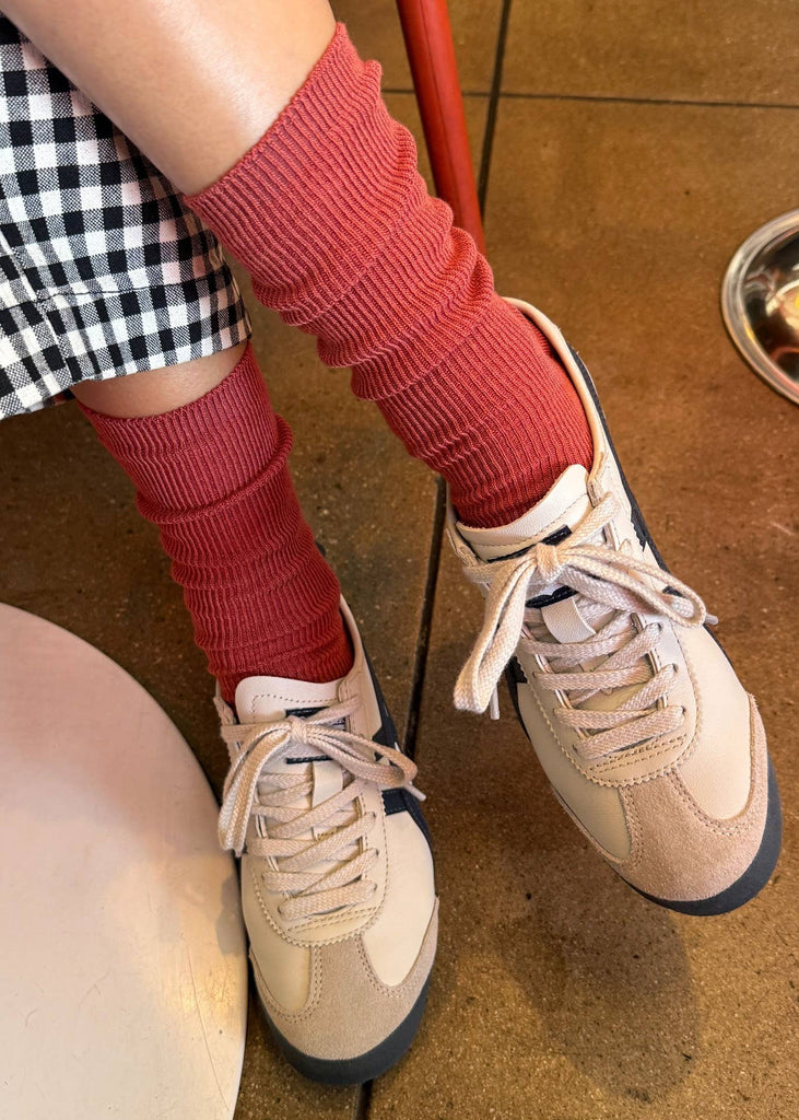 Person wearing red leg warmers and beige sneakers on a brown floor.